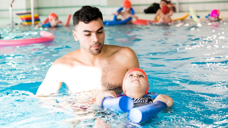 Mann unterstützt ein Kind mit Schwimmhilfen im Wasser eines Schwimmbads, während andere Schwimmer im Hintergrund sind.