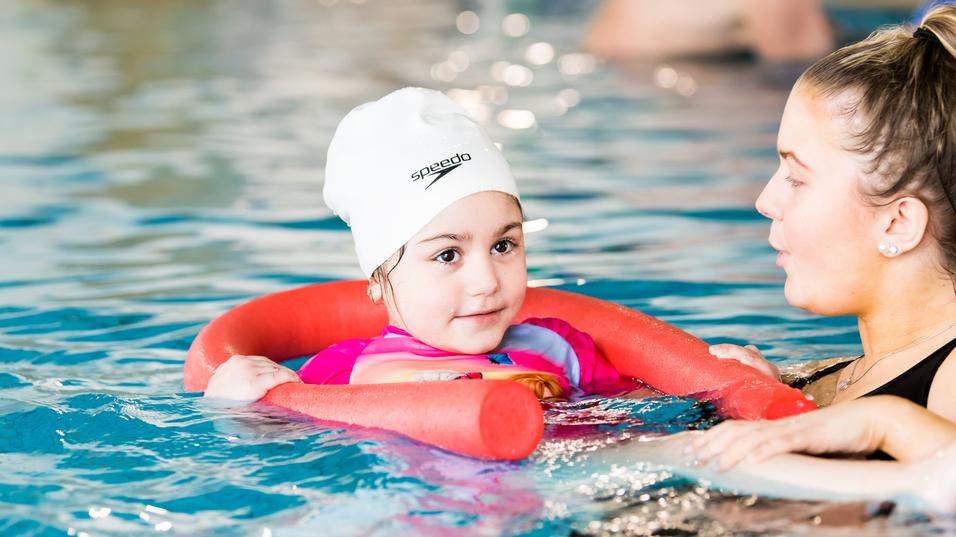 Kind mit Schwimmhilfe im Wasser, unterhält sich mit einer Erwachsenen, beide lächeln. Hintergründe zeigen andere Schwimmer.