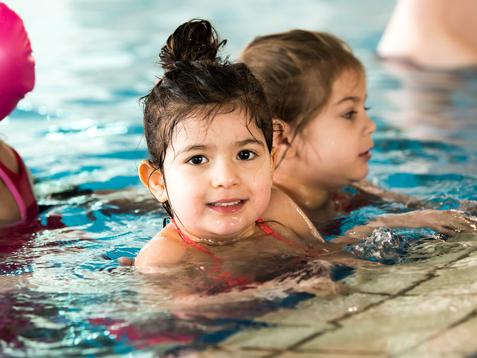 Lächelndes Mädchen mit nassen Haaren im Wasser, im Hintergrund ein weiteres Kind in der Schwimmhalle.