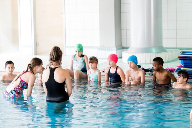 Gruppe von Kindern in Badesachen sitzt im Wasser und hört einer Trainerin am Beckenrand aufmerksam zu.