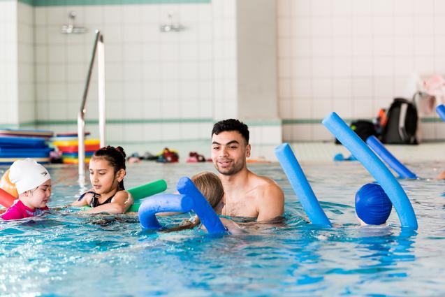 Ein Schwimmlehrer mit Kindern im Wasser, die Schwimmnudeln und Spielzeuge verwenden. Hintergrund mit Fliesen und Schwimmrequisiten.